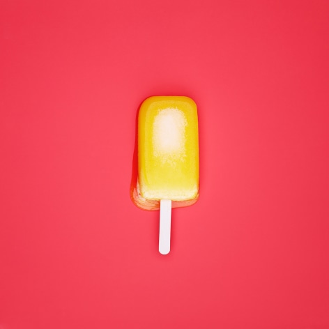 Color photo of an orange popsicle melting on a red background.