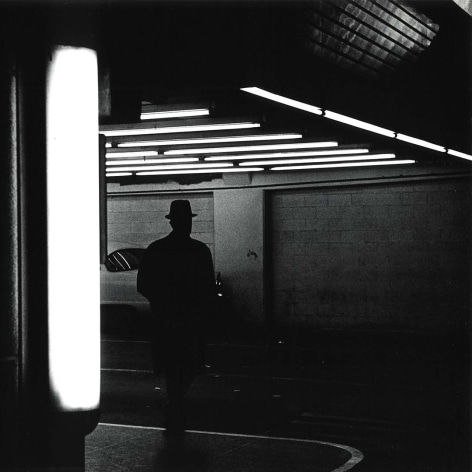 Blacand white photo of silhouetted man in a deform walking in a parking garage under florescent lights. 