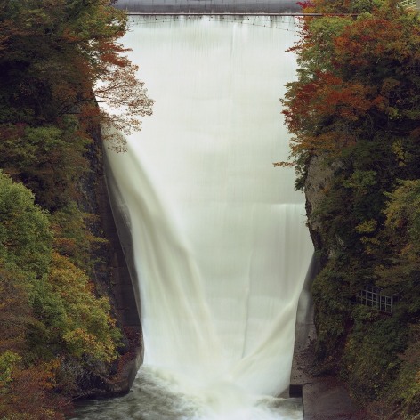 Color photo of water falling down a spillway in a forest area, with trees displaying autumn colors.
