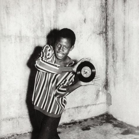 Black and white photo of an African man in a striped shirt posting and smiling with a 45 inch 1960s RCA 7-inch record single.