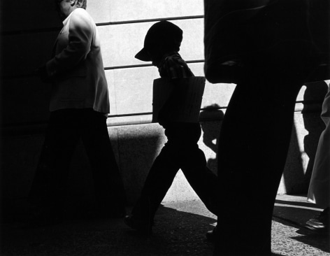 Starkly lit black and white photo of a young boy in a cap walking on a city sidewalk with his head down.