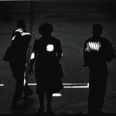 Black and white photo of pedestrians standing in shadow with spots of light falling on them.