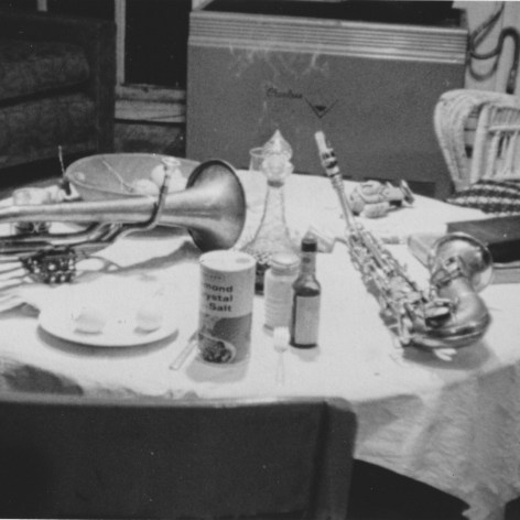 Black and white photo of an alto saxophone and a tenor horn on a kitchen table with a canister of salt and other condiments.