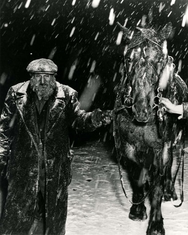 Black and white photo of a bearded street vendor, leading his horse through the snow.
