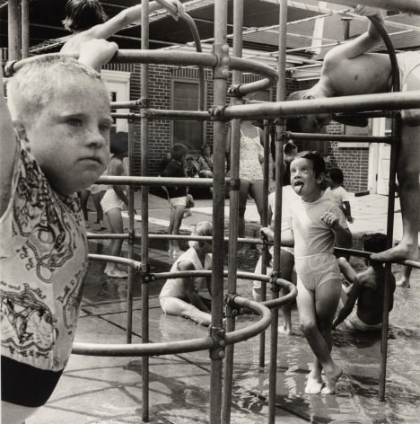 Black and white photo of kids playing on a jungle gym, a pensive boy is in the foreground while a girl in the background sticks out her tongue.