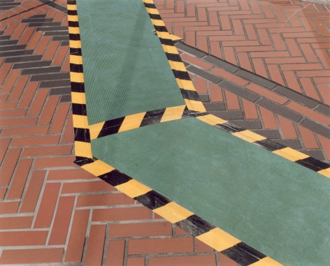Color photo of an outdoor area paved with chevron patterned brickwork, interrupted by a temporary walkway that is bounded with yellow and black striped caution tape.