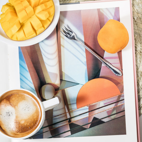 A cup of coffee, a sliced melon, and peach on top of an open photo book, showing a work by Barbara Kasten.