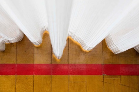 Downward facing color photo showing white curtains backlit with sun, in front of a line of red tape on a wood floor.