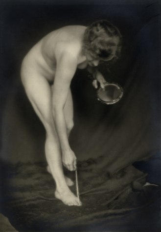 Black and white photo of nude female model, bent over, holding a hand mirror and dangling a string of pearls over her toes.
