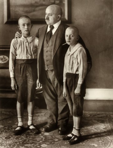 Black and white photo of a father with his tow son on either side, his hands on their shoulder and back, all with shaved heads.