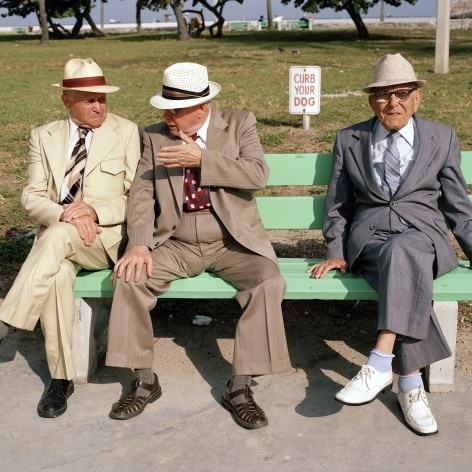 Color photo of three old men in suits and hats sitting on a bench, two talk and one faces the camera.