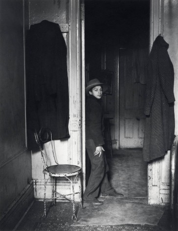 Black and white photo of a boy wearing a hat, standing in the middle of a room, looking at the camera as he leans against an entryway.