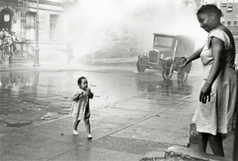 Black and white photo of a young child walking away crying with anan open fire hydrant behind her, her mother reaches out her hand to welcome her back.