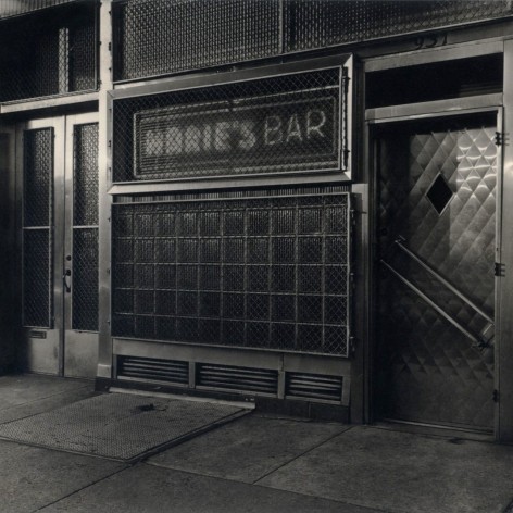 Black and white of a neighborhood bar with windows covered in protective e metal grating, a neon sign says "Marie's Bar".
