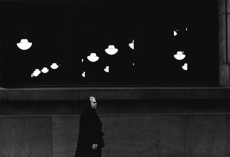 Black and white photo of a white haired woman walking on a sidewalk in front of a building with suspended lights inside.