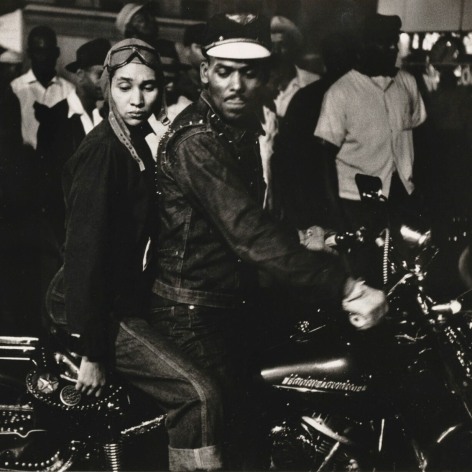 Black and white photo of two Black motorcycle riders, a man and a woman, on a large motorcycle with their eyes downcast, in front of a crowd of Black onlookers.