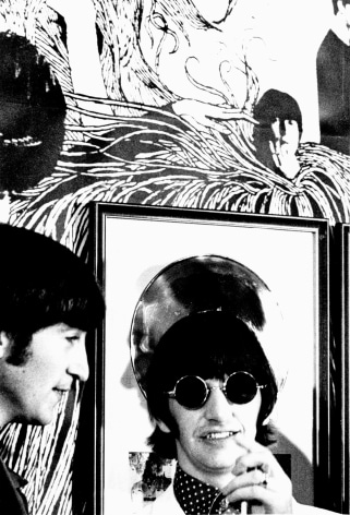 Black and white image from a Beatles press conference in 1966. Close-up of John Lennon  in front of enlargement of Revolver album art and a framed gold record..