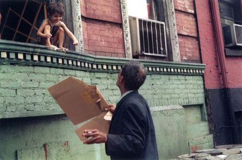 Photo of an old man holding a discarded cardboard box, looking up at a young child perched in a windowsill and laughing.