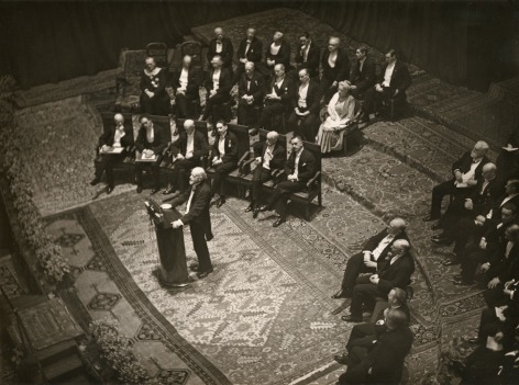Black and white photo showing the ceremony to present Thomas Mann the Nobel Prize in literature. A man speaks at the podium, with guests seats behind.