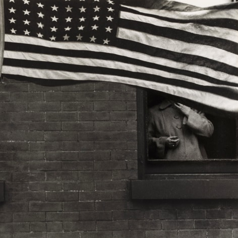 Black and white photo of a person looking out of a brick apartment building's window, their face totlaly obscured by the shadow of a waving American flag.