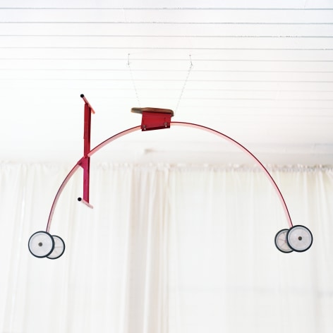Colro photo of an oddly shaped red four wheel cycle hanging from the ceiling in a white room.