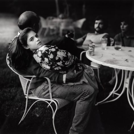 Black and white photo of an adolescent girl sitting on an adults lap at an outdoor table, looking back at the camera.
