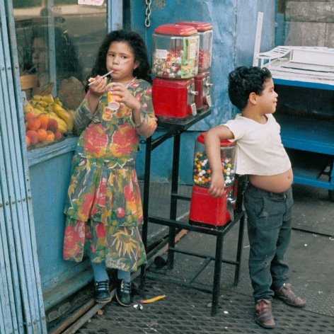 Color photo of two children hanging out by a gumball machine outside a small shop.