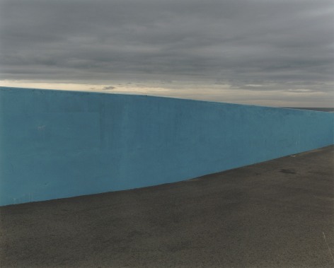 Color photo of a blue wall running along a beach, beneath a cloudy sky, the ocean can be glimpsed on the other side of the wall, on the right.