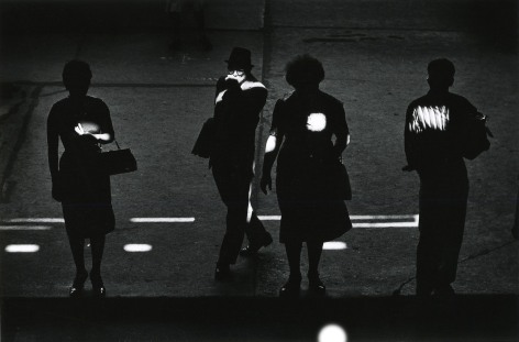 Shadowy black and white photo showing pedestrian in shadow, with spots of light on them.