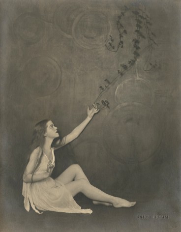 Staged black and white photo of a seated young woman in a white dress, reaching up towards a dangling vine. The background shows decoratively painted swirls and whorls.