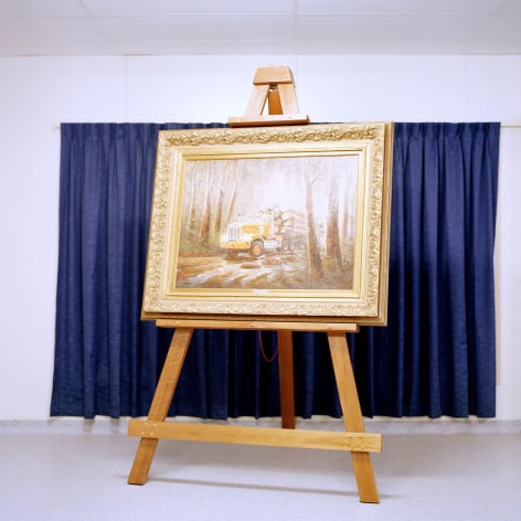 Color photo of a framed painting of a logging truck on an easel, in front of blue curtains.