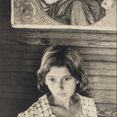 Early 20th entry photographic portrait of a young girl in. applaud dress with downcast eyes, in front of a clapboard was with a print of the Madonna and Child on it.