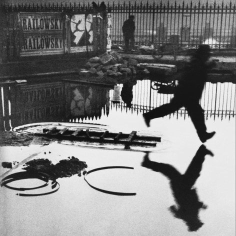 Black and white photo of a man bounding over a flooded plaza, reflected in the water below.