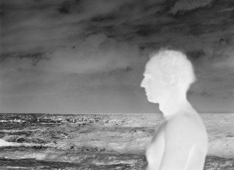 Black and white negative photograph of a shirtless man, seen in profile by the ocean shore.
