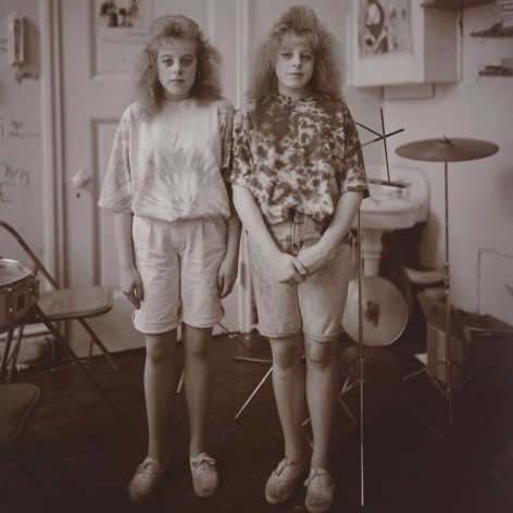 Posed black and white photo of twins sisters in a room with a drum kit.