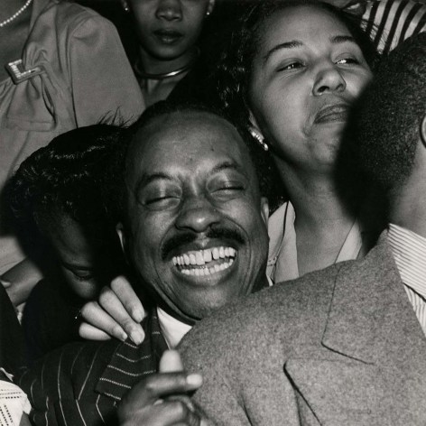 Vintage black and white photo of an elated Black man, dressed in a suit in a crowded jazz club. People are shoulder too shoulder.