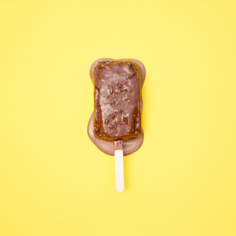 Color photo of a chocolate popsicle melting on a yellow background.