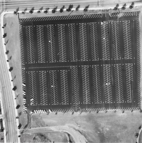 Aerial black and white photo of large, mostly empty, parking lot, showing the panted pattern created by the parking spots.