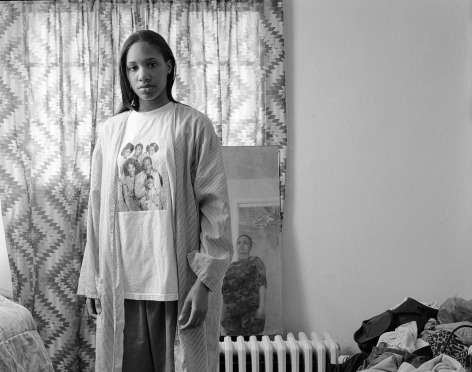 Black and white photo portrait of young Black woman,  in a domestic interior, looking at the camera, wearing a faded Cosby Show t-shirt. An older woman across the room can be seen in the mirror next to her.