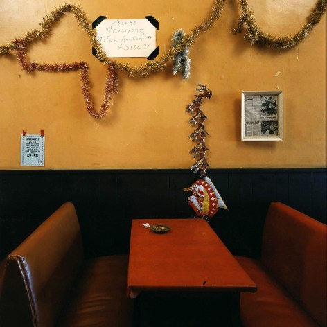 Color photo of a dilapidated red restaurant booth with a red tabletop, orange wall, and Chsitmaas tinsel and decor haphazardly affixed to the wall, as well as some taped and framed signs.