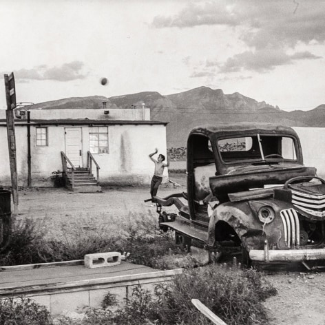Black and white photo of a shirtless boy shooting a basketball in front of a house in a desert landscape with a broken down car in the foreground.