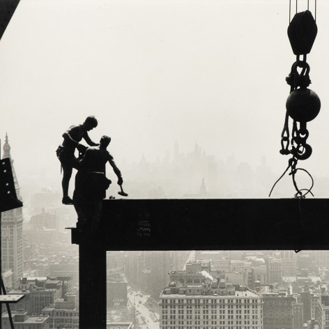 Black and white photo showing silhouette of workers hammering beams on Empire State Building with New York City skyline in the background