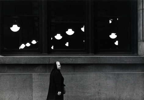 High contrast black and white photo of an old woman in a he'd scarf walking on a city sidewalk in front of a large window showing a number of mid-century half-globe hanging  light fixtures.