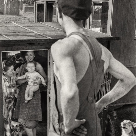 Black and white photo of a man with his hands on his hips looking at two women holding a baby, on a houseboat, while two dogs look on.