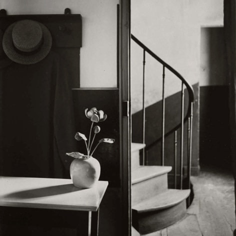 Black and white photo of a flower vase on a table in front of a coat and hack on a rack, with a spiral staircase behind.