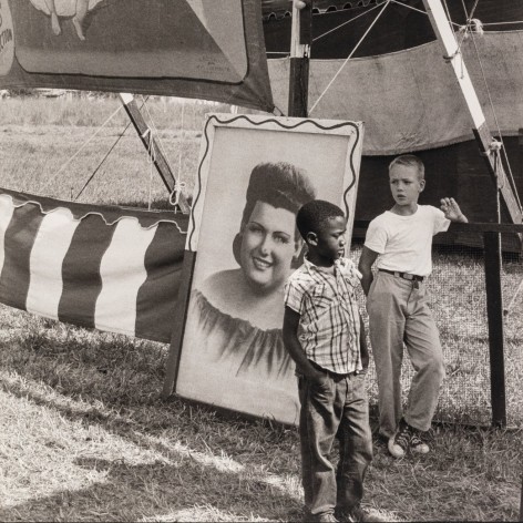 Black and white photo of young boys, one back and one white, standing in front of a circus sideshow, with a sign advertising a "2 HEADED BABY"