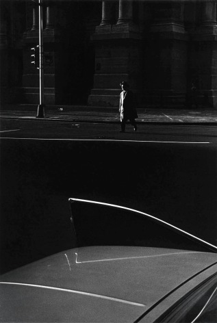 Black and white photo of a man in trenchant and defer crossing street, 1960's vintage car with tail fins is in the foreground.
