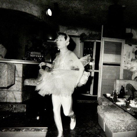 Black and white photo of a cross dressing man, wearing a ballerina outfit, parading in front of Japanese bar patrons who are seated and watching.