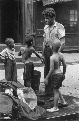 Black and white photo showing a NYC street scene in the 1940s, with a group of young boys gathered around ice that a tall ice man has just delivered on the sidewalk.