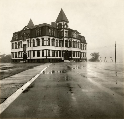 Black and white phto of a large Victorian building in front of a schoolyard that is wet with rain. 